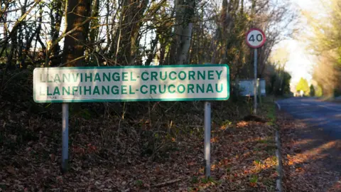 A sign for the village of Llanvihangel Crucorney on a leafy roadside verge. Behind it are trees and a 40mph sign.