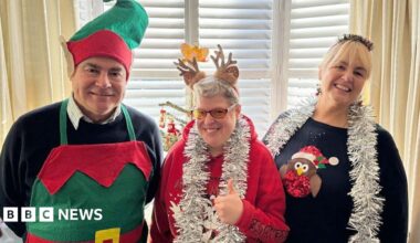 Left to right: Mike, Clare and Kim. Mike is wearing an elves apron and hat. Clare has a red Christmas jumper with silver tinsel round her neck and reindeer ears. Kim with blonde hair wears a navy blue Christmas jumper and silver tinsel around her neck. They are all standing in front of a Christmas tree and smiling.