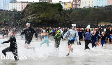 A large crowd of people, some in fancy or Christmas dress, is running into the sea. There is a large moss-covered rock on the beach behind them and a raised walkway around the beach with people standing spectating. A grassy cliff frames the beach with a street above it, backed by blocks of brightly painted townhouses in different colours.