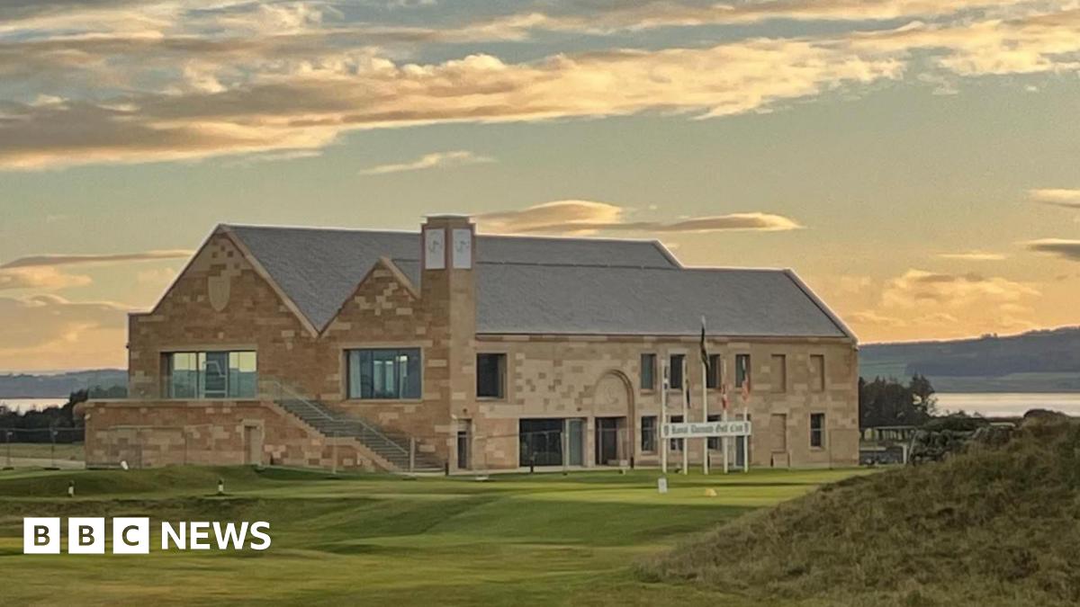 The sun has just gone down and clouds are lit in orange and gold colours above the new clubhouse at Royal Dornoch. It is a large building made of sandstone with a slate roof. A golf tee point is located in front of the building.