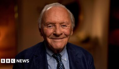 Sir Anthony Hopkins looking to camera and smiling; dressed in navy blue suit, light blue shirt, and dark blue and white tie