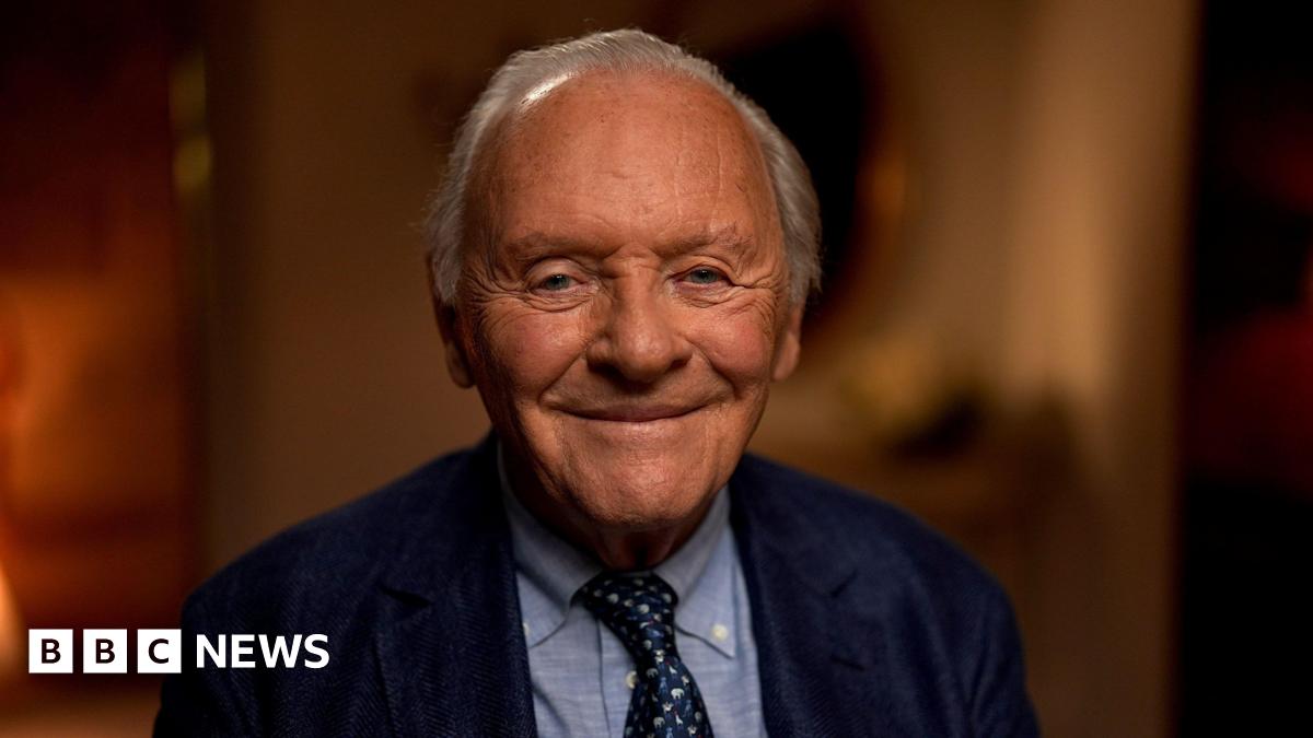 Sir Anthony Hopkins looking to camera and smiling; dressed in navy blue suit, light blue shirt, and dark blue and white tie