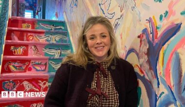 A blonde women smiles in front of a colourful staircase in the hallway of her family home. Painted birds cover the walls.