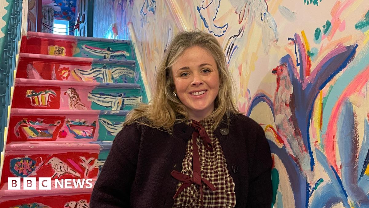 A blonde women smiles in front of a colourful staircase in the hallway of her family home. Painted birds cover the walls.