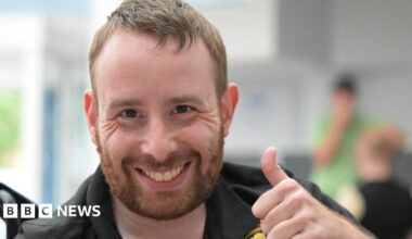 A man with short light brown hair is giving a thumbs-up gesture while seated indoors. He is wearing a black polo shirt with gold embroidered logos, including one circular badge on the chest. The background is softly blurred, showing another person in green.