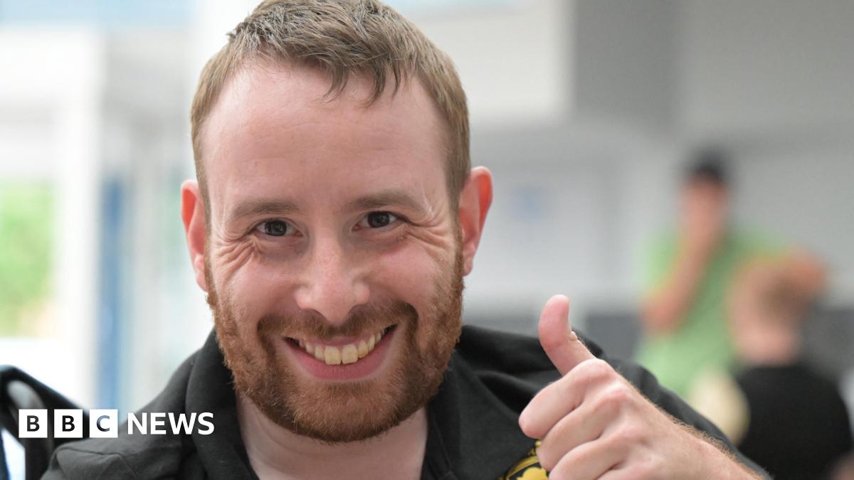 A man with short light brown hair is giving a thumbs-up gesture while seated indoors. He is wearing a black polo shirt with gold embroidered logos, including one circular badge on the chest. The background is softly blurred, showing another person in green.