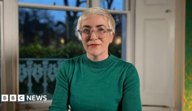 Alice Sullivan sits in a room looking at the camera. She has short platinum blonde hair, glasses and bright lipstick. She's wearing an emerald green jumper.