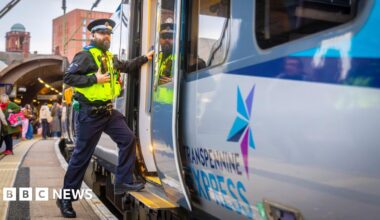 A man in a Police Community Support Officer uniform steps on to a train from a busy platform. The train door is open. A sign on the side of the train says TransPennine Express.