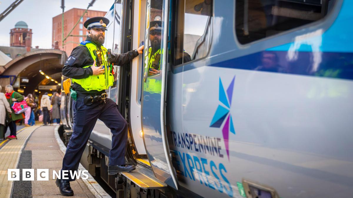 A man in a Police Community Support Officer uniform steps on to a train from a busy platform. The train door is open. A sign on the side of the train says TransPennine Express.