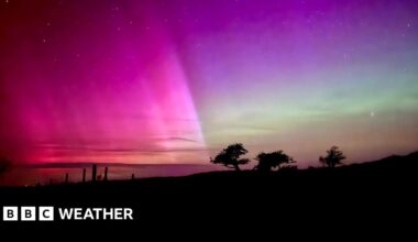A photo of the aurora borealis in Dorset showing shades of green and pink dancing across the night sky with a silhouetted landscape in the foreground