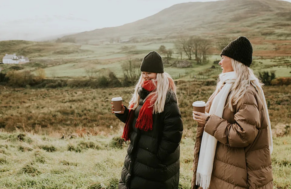 Two beautiful, stylish young woman walk through the countryside, side-by-side, clutching takeaway coffee cups. They have a lighthearted chat, enjoying the company. They are bundled up in warm clothing to reflect the season, which includes padded jackets, wooly hats, and large scarves.