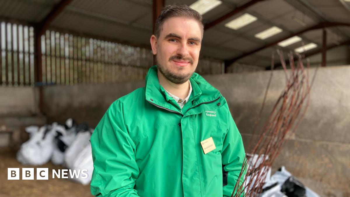 A man with brown hair and facial hair smiles as he stands in a barn, holding a sapling. Large white bags containing saplings can be seen behind him. He is wearing a green, Forestry England branded coat, and a name badge.