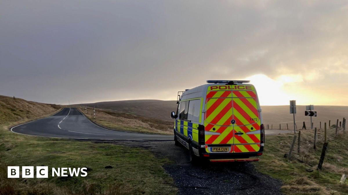 A police van parked on a path of a bending road. The sun is setting over the hills in the distance.