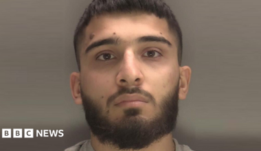 A close-up of a man's face. He has a moustache and beard and is looking straight at the camera, and is pictured against a grey background