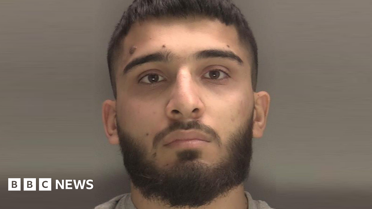 A close-up of a man's face. He has a moustache and beard and is looking straight at the camera, and is pictured against a grey background