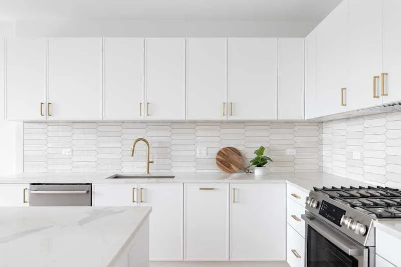 A beautiful kitchen detail with white cabinets, a gold faucet, white marble countertops, and a brown picket ceramic tile backsplash.