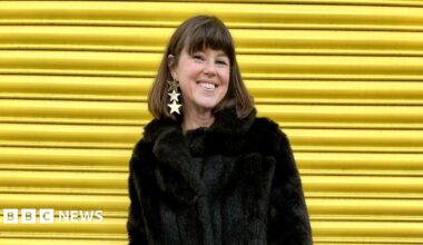 Caroline Jones is standing against a bright yellow background. She wears a black faux-fur coat. She has dark brown shoulder-length straight hair and a fringe and you can see a large drop earring made up of three gold stars in her right ear. She is smiling broadly at the camera.