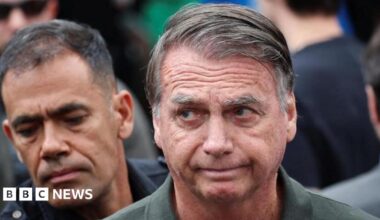 Brazil's former president Jair Bolsonaro is captured mid-wave, his fingers just visible, looking into the distance. His hair is combed over to the right and he is wearing a green khaki shirt.
