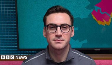 A man is looking straight at the camera. He has short jet black hair, which is slicked back. He has black-rimmed glasses. He is wearing a grey sports top with a zip. He is sitting in a podcast studio with a pink sofa, behind his head is a TV with the logo for The State of Us podcast, which is a teal background with a stylised image of Northern Ireland in pink.