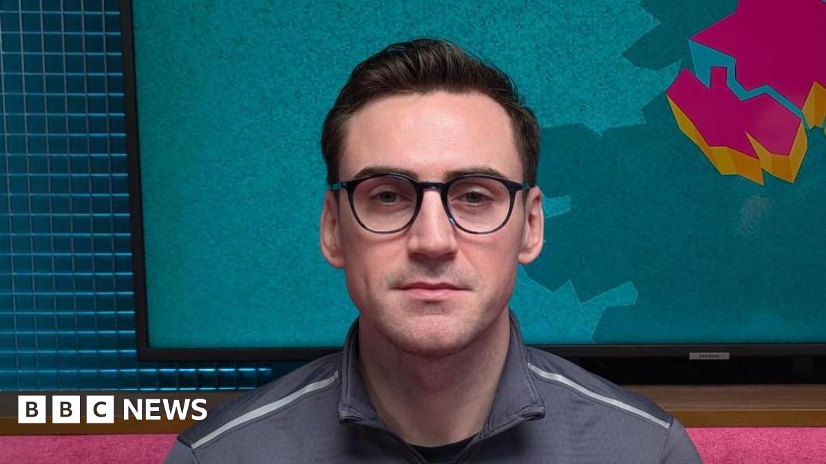 A man is looking straight at the camera. He has short jet black hair, which is slicked back. He has black-rimmed glasses. He is wearing a grey sports top with a zip. He is sitting in a podcast studio with a pink sofa, behind his head is a TV with the logo for The State of Us podcast, which is a teal background with a stylised image of Northern Ireland in pink.