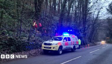 A roadside cave rescue operation under way, with a specialist vehicle on blue lights next to a wooded bank