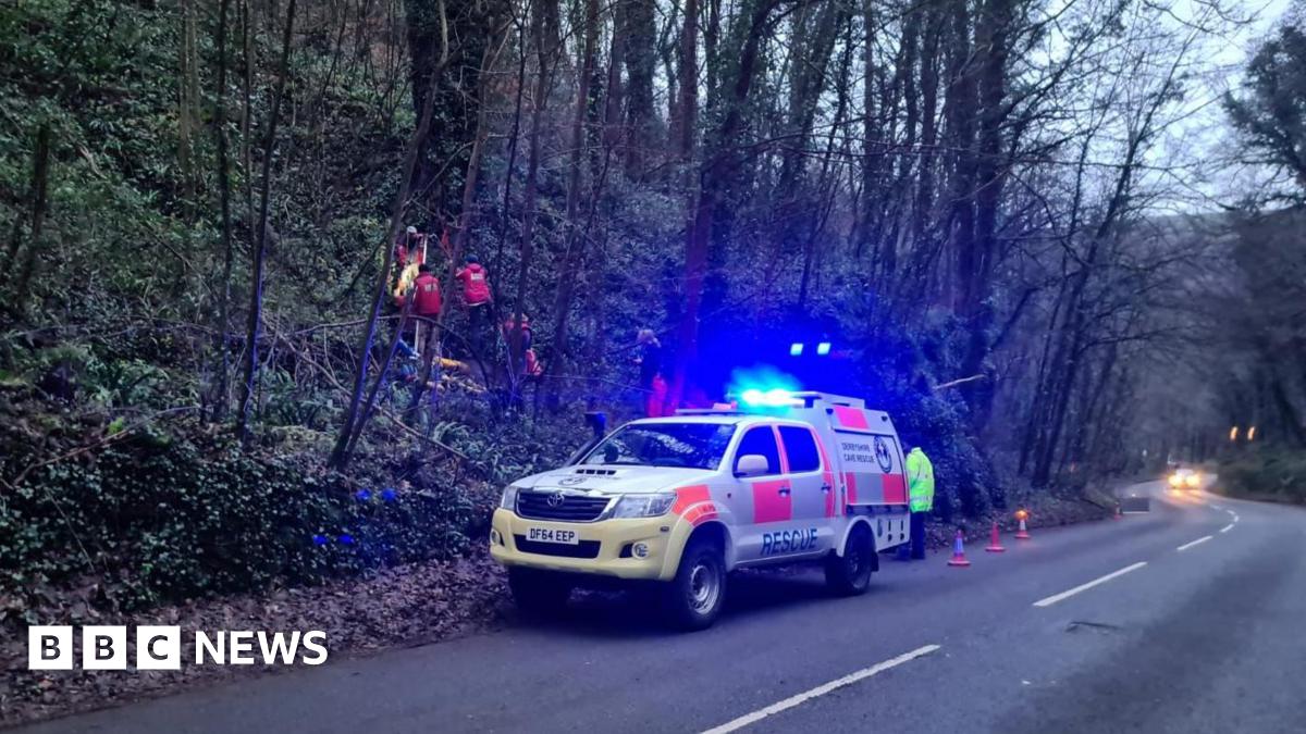 A roadside cave rescue operation under way, with a specialist vehicle on blue lights next to a wooded bank