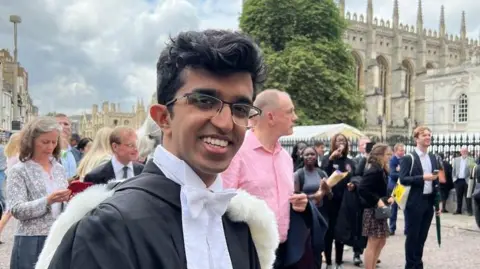 Ray Amjad Ray wearing a graduation gown in front of one of the historic colleges in Cambridge. He has glasses and is smiling at the camera. Behind him 