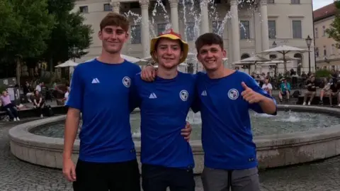 Handout The three young men are wearing blue football jerseys, posing and smiling at the camera in front of a large fountain in a public space.