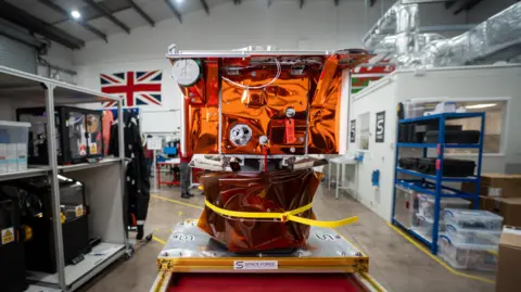 Tony Jolliffe/BBC News A life-size mock up of the space factory satellite sitting on a workbench inside Space Forge's Cardiff headquarters. It is square-shaped, about the size of a microwave, and is covered in reflective orange foil with various smaller components attached. The workshop in the background has high ceilings and has shelves filled with equipment and storage boxes. A large Union Jack flag hangs on the wall.