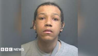 A woman police officer wearing a yellow coat stands with folded arms outside a fast food shop. Kneeling to her right is a crime scene investigator dressed in a white suit.