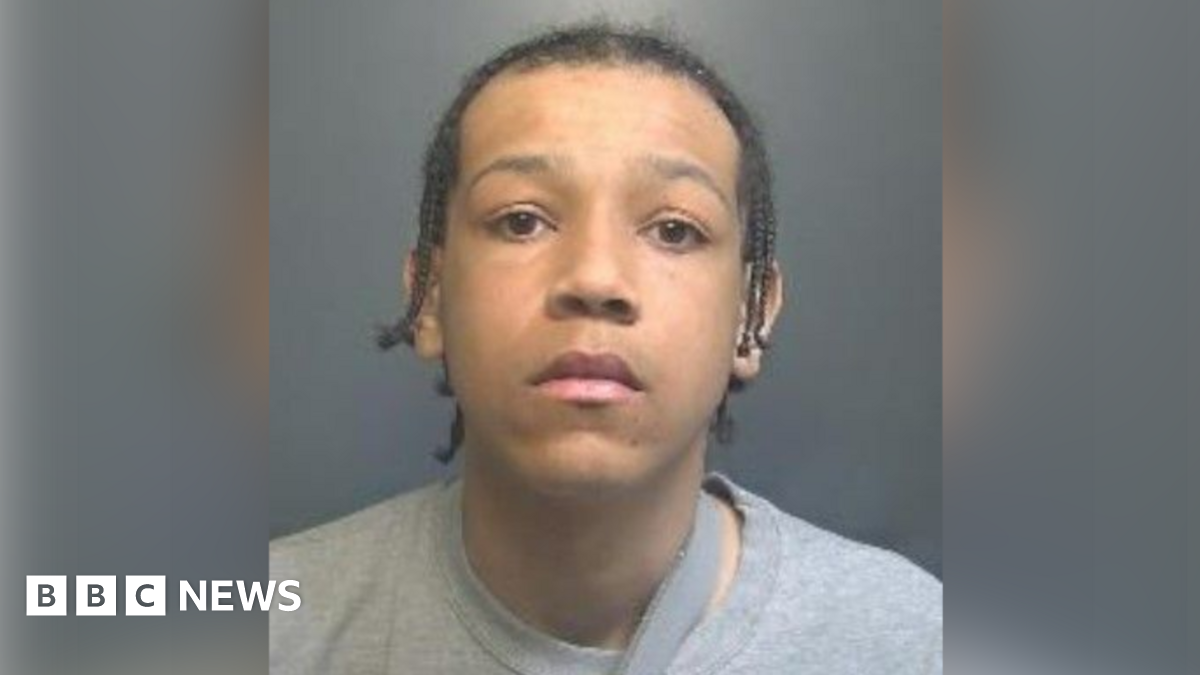A woman police officer wearing a yellow coat stands with folded arms outside a fast food shop. Kneeling to her right is a crime scene investigator dressed in a white suit.