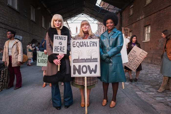 Helen George as Trixie Franklin, Natalie Quarry as Rosalind Clifford and Renee Bailey as Joyce Highland, holding signs saying VENUS IS HERE and EQUAL PAY NOW.