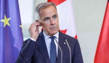 Government official at a podium adjusts an earpiece, with EU, Canada and Germany flags behind him.