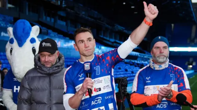 Three men stand in a football stadium. Two of them are wearing blue, red, and white sports gear and another is wearing a grey puffer jacket and black cap.