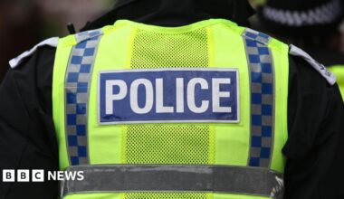 The back of a police vest. It is neon yellow and has two vertical stripes in navy and grey chequers. A navy blue rectangle says "police" in white lettering.