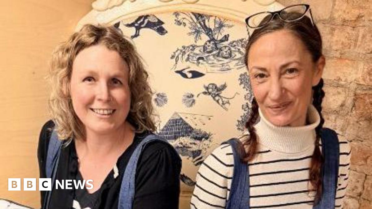 Karen Evans, with blonde curly hair, wearing a black top, blue tights and black boots, holding a cushion with the artwork on. The cushion is white and has blue illustrations on it. Sitting next to her is Maddy Humberstone, holding a similar cushion, with brown hair tied back, wearing jeans and a white stripey top and Converse trainers.