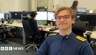 A man wearing a navy blue top with yellow shoulder pads. He is sat in an office with people and computers in the background.