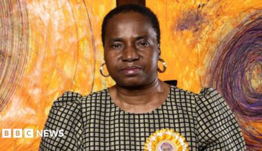 A brown NC500 sign post which says it is 27 miles to Ullapool next to a country road with hills in the background.