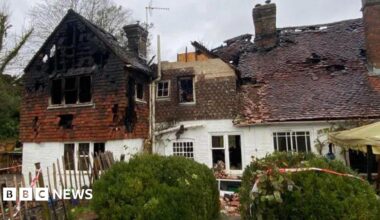 A house that has burned down, with holes in the roof and red and white safety tape around it and on the bushes at the front of the house. The sky in the background is grey and bushes can be seen on the left-hand-side.