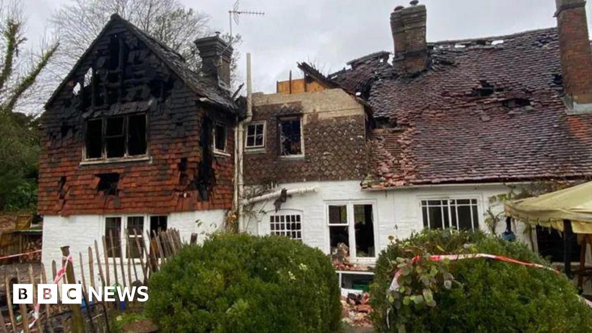 A house that has burned down, with holes in the roof and red and white safety tape around it and on the bushes at the front of the house. The sky in the background is grey and bushes can be seen on the left-hand-side.