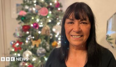 A woman with shoulder length dark hair, wearing a black top standing in front of a Christmas tree.