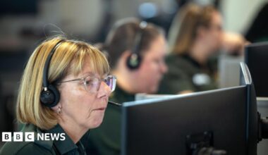 A blonde woman wearing glasses and a green polo shirt. She is sitting inside an ambulance call room with several others.
