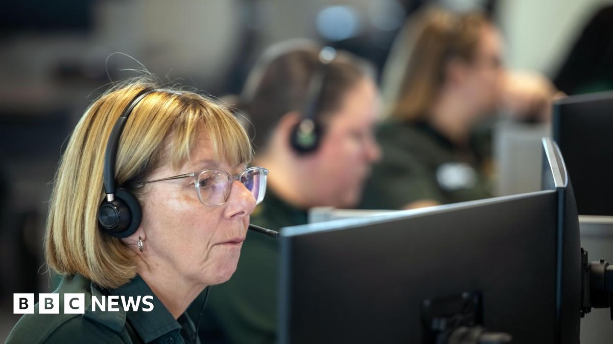 A blonde woman wearing glasses and a green polo shirt. She is sitting inside an ambulance call room with several others.
