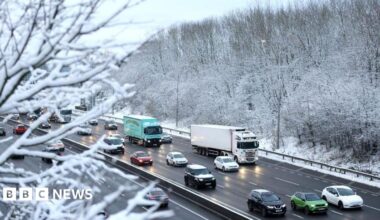 Cars and lorries travel along the M62 near Bradford on a wintry day. Headlights reflect on the wet carriageway and the picture is framed by snow-covered branches.