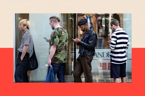 Getty Images Four men stand in a queue, looking at their smartphones.