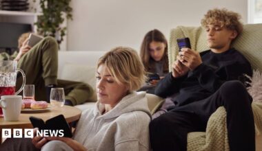 A family (mother, two children, father) sitting down in the living room - on a sofa and the floor - all looking at their phones separately and looking unimpressed.