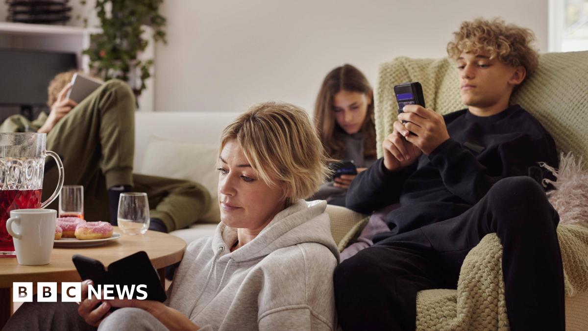 A family (mother, two children, father) sitting down in the living room - on a sofa and the floor - all looking at their phones separately and looking unimpressed.
