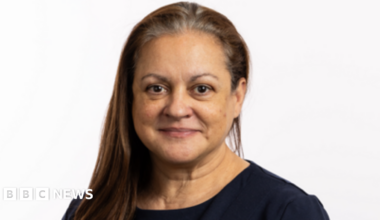 A woman with long dark hair, swept forward over one shoulder, is looking at the camera. She is wearing a dark top and is smiling