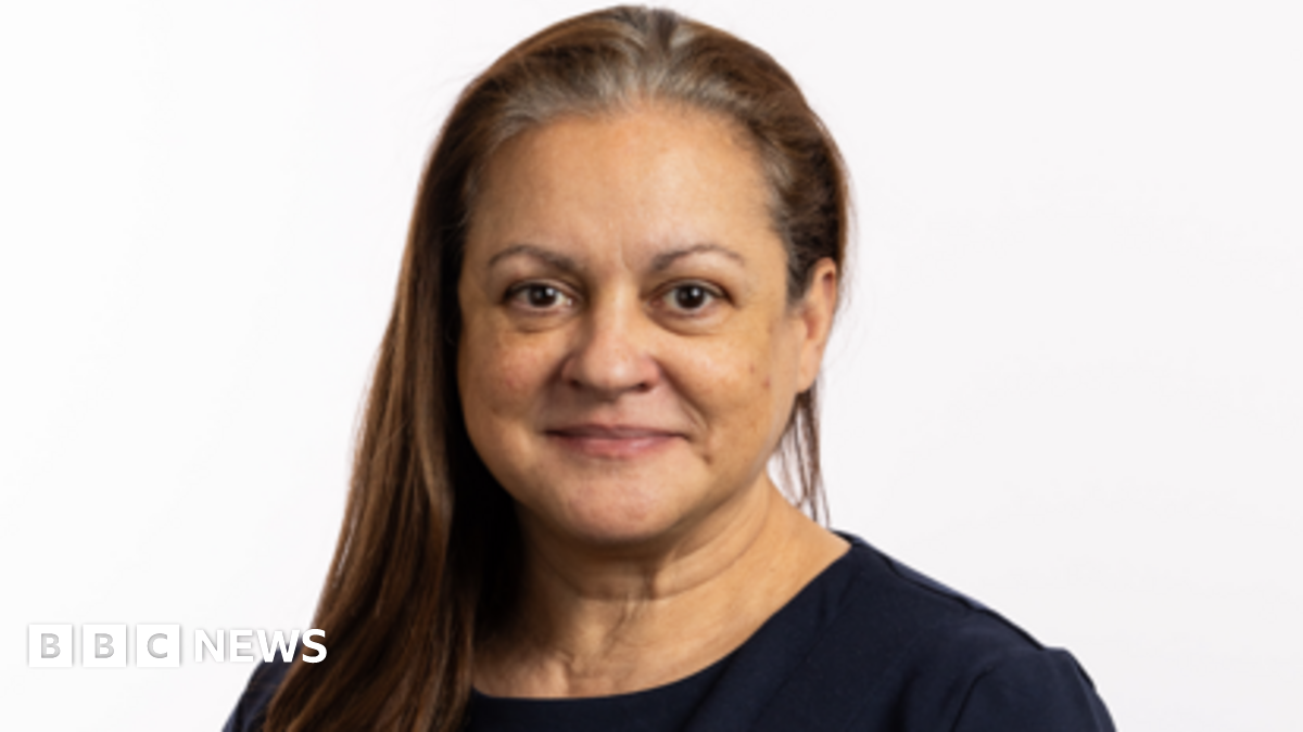 A woman with long dark hair, swept forward over one shoulder, is looking at the camera. She is wearing a dark top and is smiling
