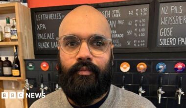 Head and shoulders image of Vinny Rosario looking straight at the camera. He is bald with a thick dark beard and moustache. He is wearing a grey sweatshirt and standing in front of a wall with lots of different coloured beer taps on it.
